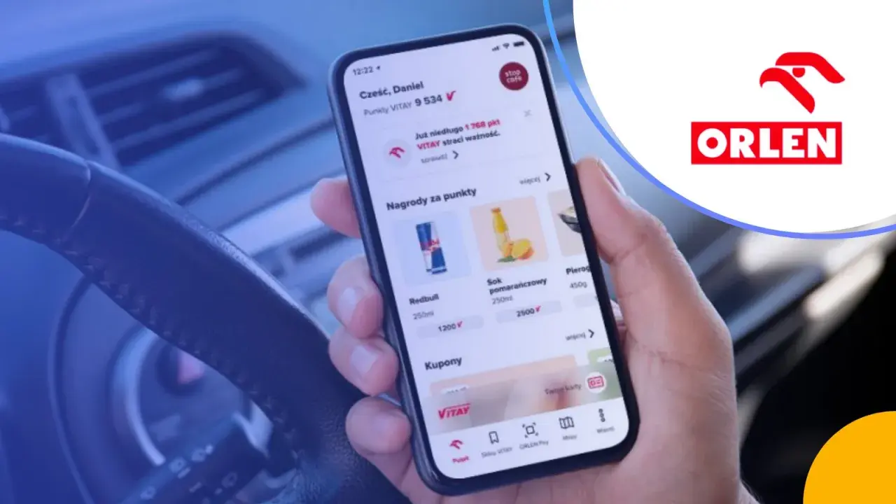 Dłoń trzyma smartfon z aplikacją ORLEN Pay, pokazującą punkty VITAY i nagrody. Kierownica samochodu w tle.
