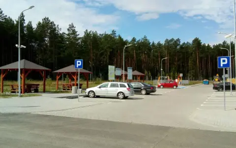 Parking przy lesie, z wiatami i ławkami. W tle las, a na parkingu kilka samochodów. To idealne miejsce na postój w drodze, może nawet w pobliżu **Orlen Sople**.