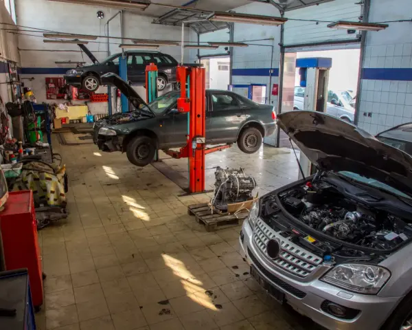 Warsztat samochodowy z kilkoma autami na podnośnikach, w tym ciemnym sedanem, który wygląda jak p1351 peugeot. Silnik wyjęty z jednego z pojazdów.
