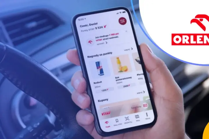 Dłoń trzyma smartfon z aplikacją ORLEN Pay, pokazującą punkty VITAY i nagrody. Kierownica samochodu w tle.